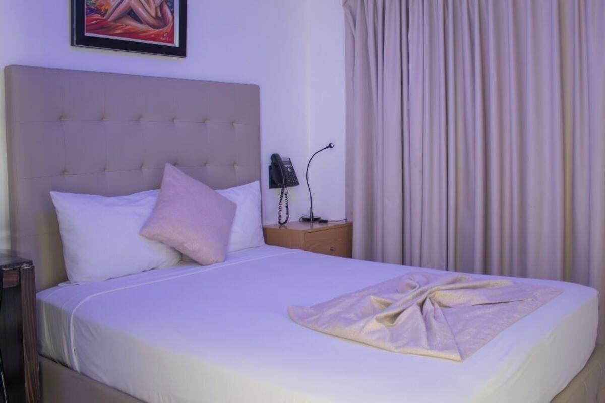 A neatly made bed with a white comforter and a folded blanket, a bedside lamp, a phone, and a framed picture on the wall.