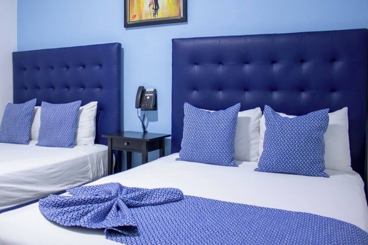 A hotel room with two beds, each with a blue headboard and white bedding. There is a small bedside table with a phone and a painting on the wall above the beds.