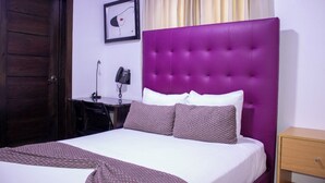 Un dormitorio con cabecera morada, ropa de cama blanca y manta marrón con estampado.