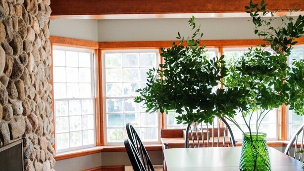 A dining area with a stone wall, wooden beams, a large window, and a green plant in a vase.