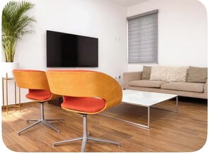 Ein modernes Wohnzimmer mit Fernseher, zwei orangefarbenen Sesseln, einem weißen Couchtisch und einer Couch.