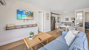 A modern living room with a sofa, coffee table, and a TV mounted on the wall. There is a kitchen area visible in the background with a refrigerator and a dining table. - The Drift (Cocoa Beach)
