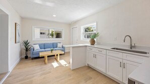 A modern kitchen with white cabinets, a sink, and a blue sofa in the adjacent living area. - The Drift (Cocoa Beach)
