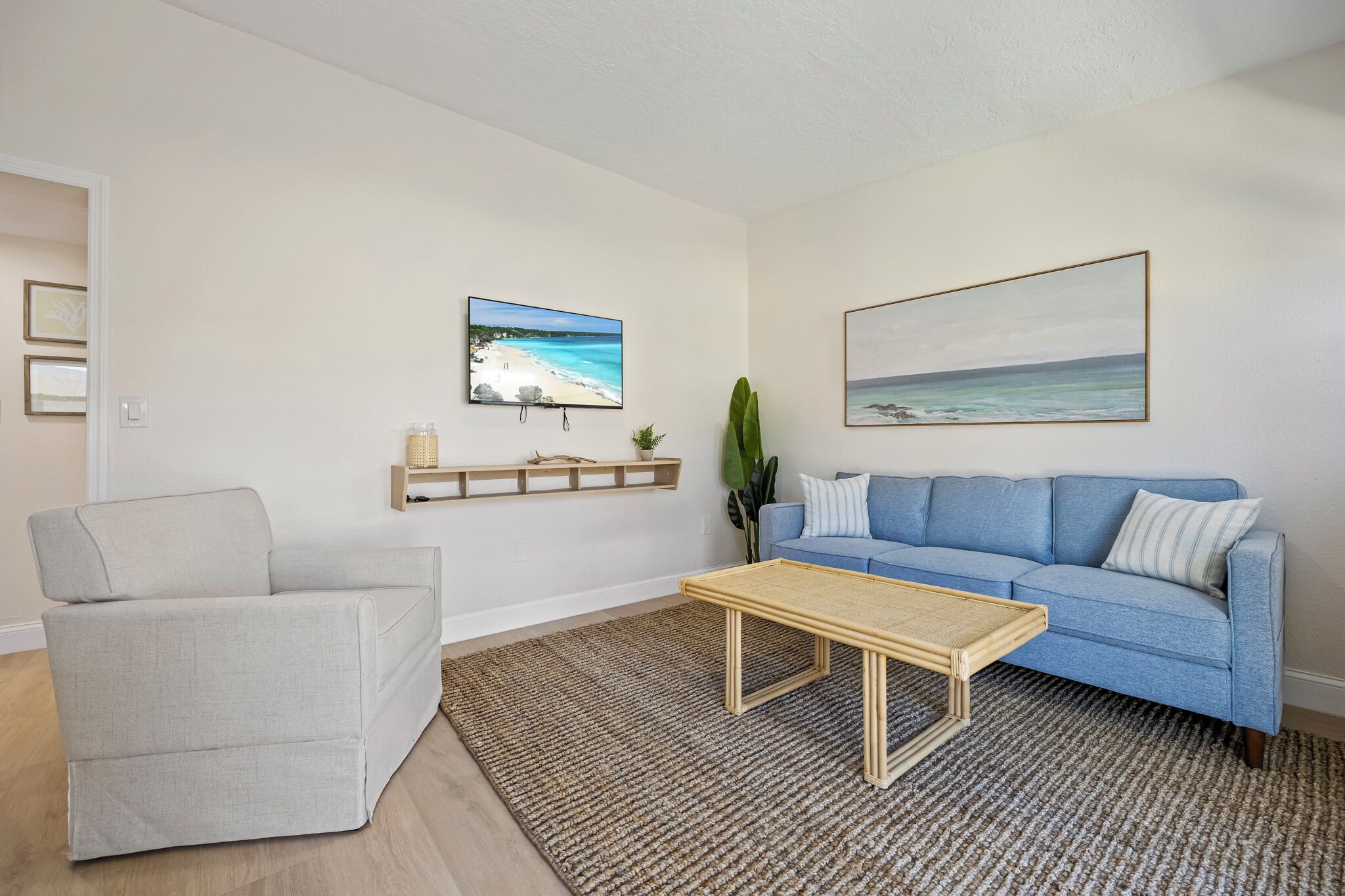 A living room with a light blue sofa, a grey armchair, and a wooden coffee table. There are two framed pictures on the wall, one of a beach and another of a landscape.