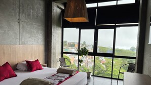 Habitación de hotel moderna con una cama grande, vistas a un paisaje urbano a través de amplios ventanales y una lámpara colgante.