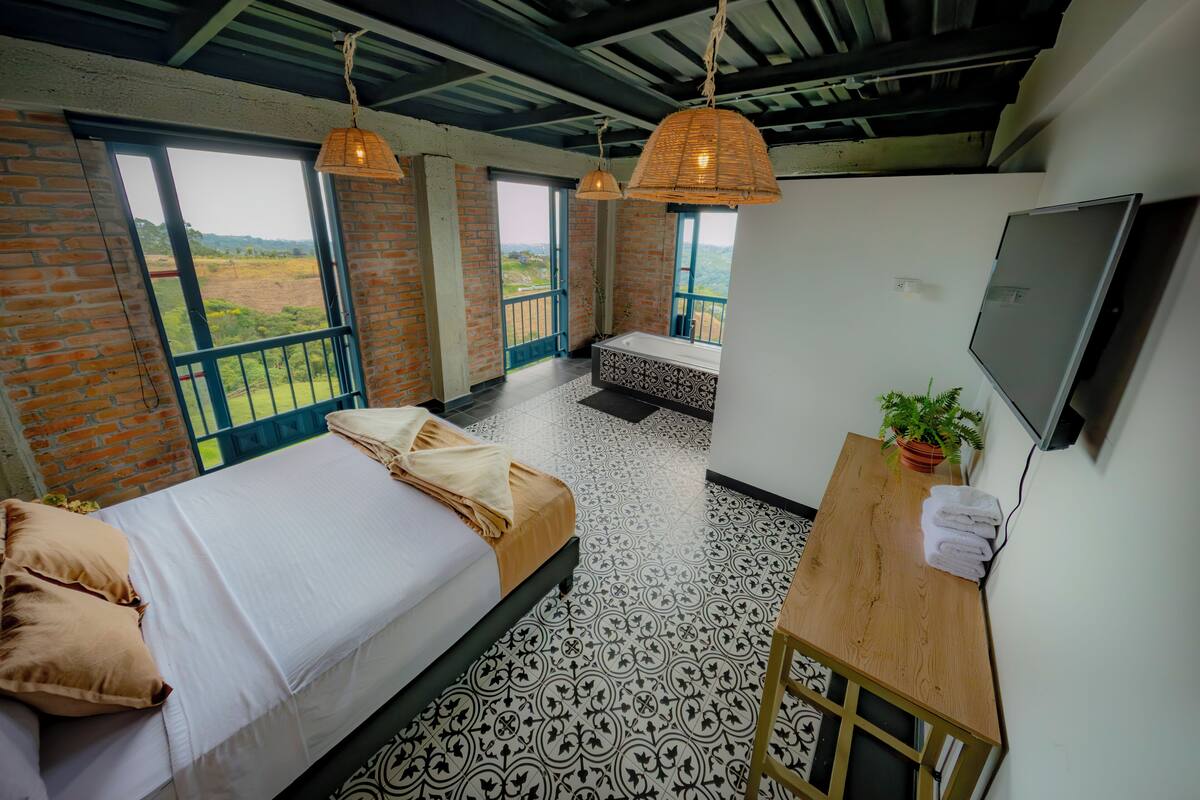 A bedroom with a bed, a wooden table, a TV, and a balcony with a view.