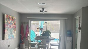 A spacious living room with a ceiling fan, a dining area with a round table and chairs, a large painting on the wall, and a view through a sliding glass door to an outdoor area. - Charming 2-bedroom pool home with AC, WiFi in Lehigh Acres (Lehigh Acres)