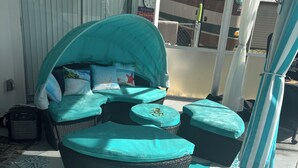 A covered outdoor seating area with a cushioned sofa and ottoman, a clock, and a sign indicating a nearby restaurant. - Charming 2-bedroom pool home with AC, WiFi in Lehigh Acres (Lehigh Acres)