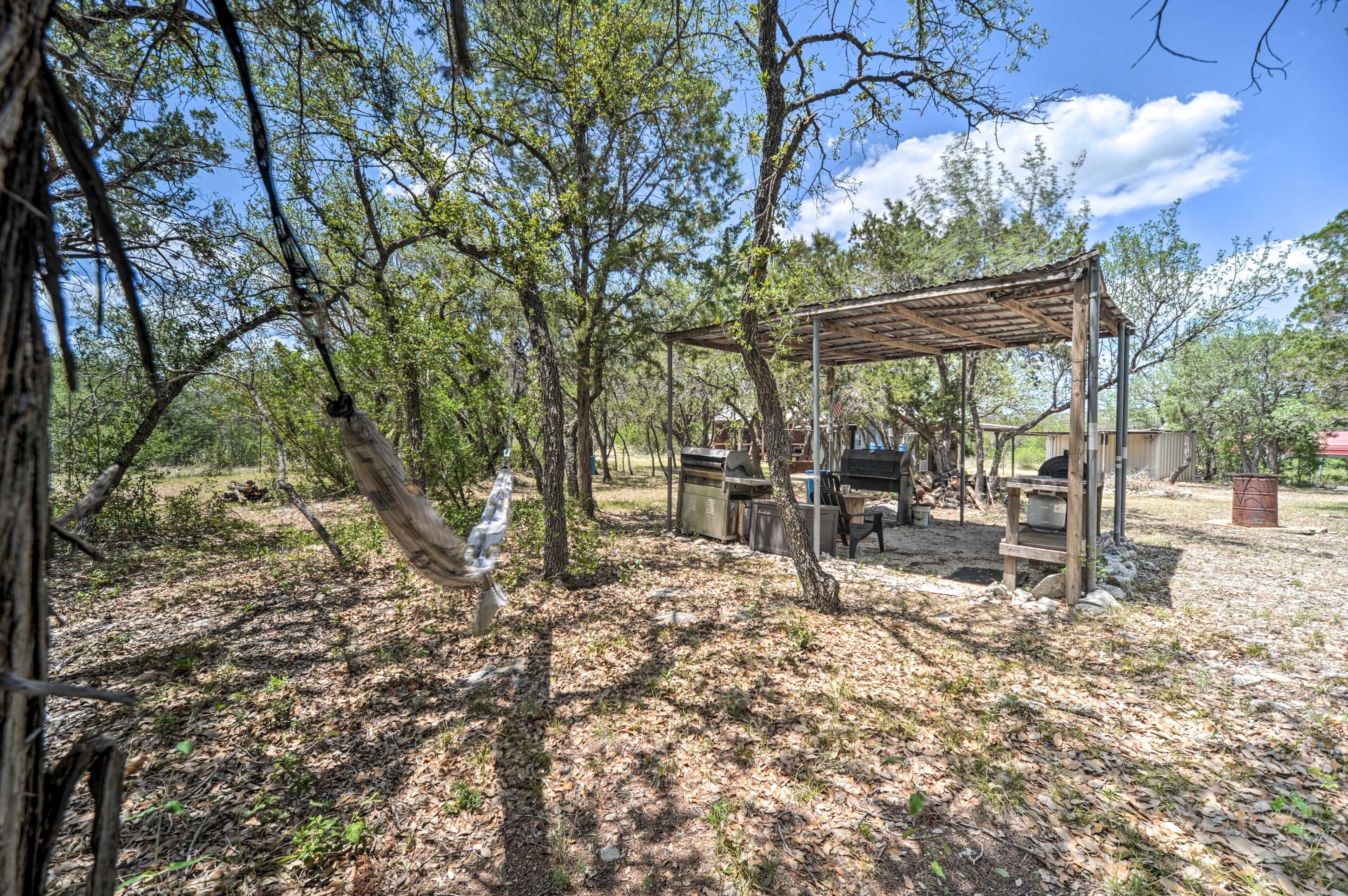 Ein Außenbereich mit einer hölzernen Pergola, Grill und Sitzgelegenheit, umgeben von Bäumen.