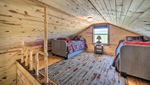 A cozy attic bedroom with wooden walls and ceiling, a window with a view, two beds, a small table, and a wooden staircase. - Rustic Cabin On 31 Acres + Near the Frio River • Pet Friendly • Sleeps 6+ (Sabinal)