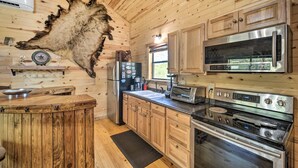 A rustic kitchen with wooden walls, a large bear hide, a refrigerator, a microwave, a stove, and a bar area. - Rustic Cabin On 31 Acres + Near the Frio River • Pet Friendly • Sleeps 6+ (Sabinal)