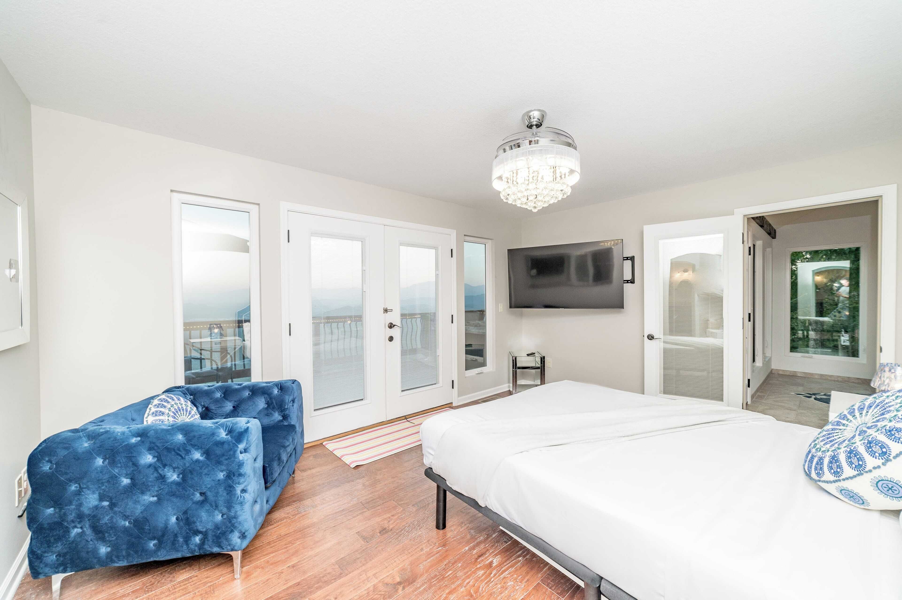A bedroom with a large bed, a blue sofa, a TV mounted on the wall, and a balcony door.