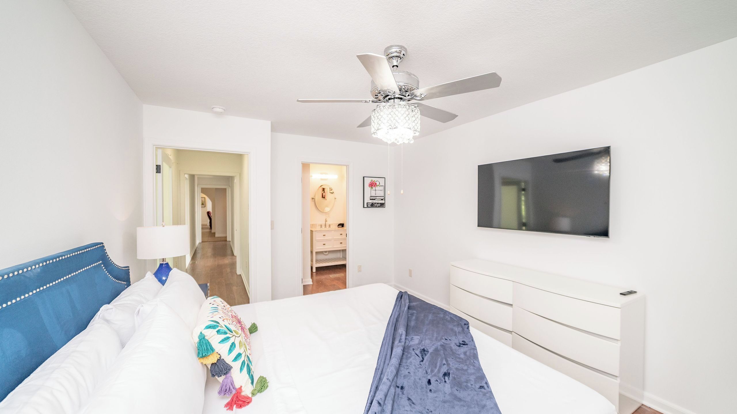 Une chambre à coucher avec un lit, un ventilateur de plafond, une télévision, une armoire et une salle de bain visible à travers une porte ouverte.