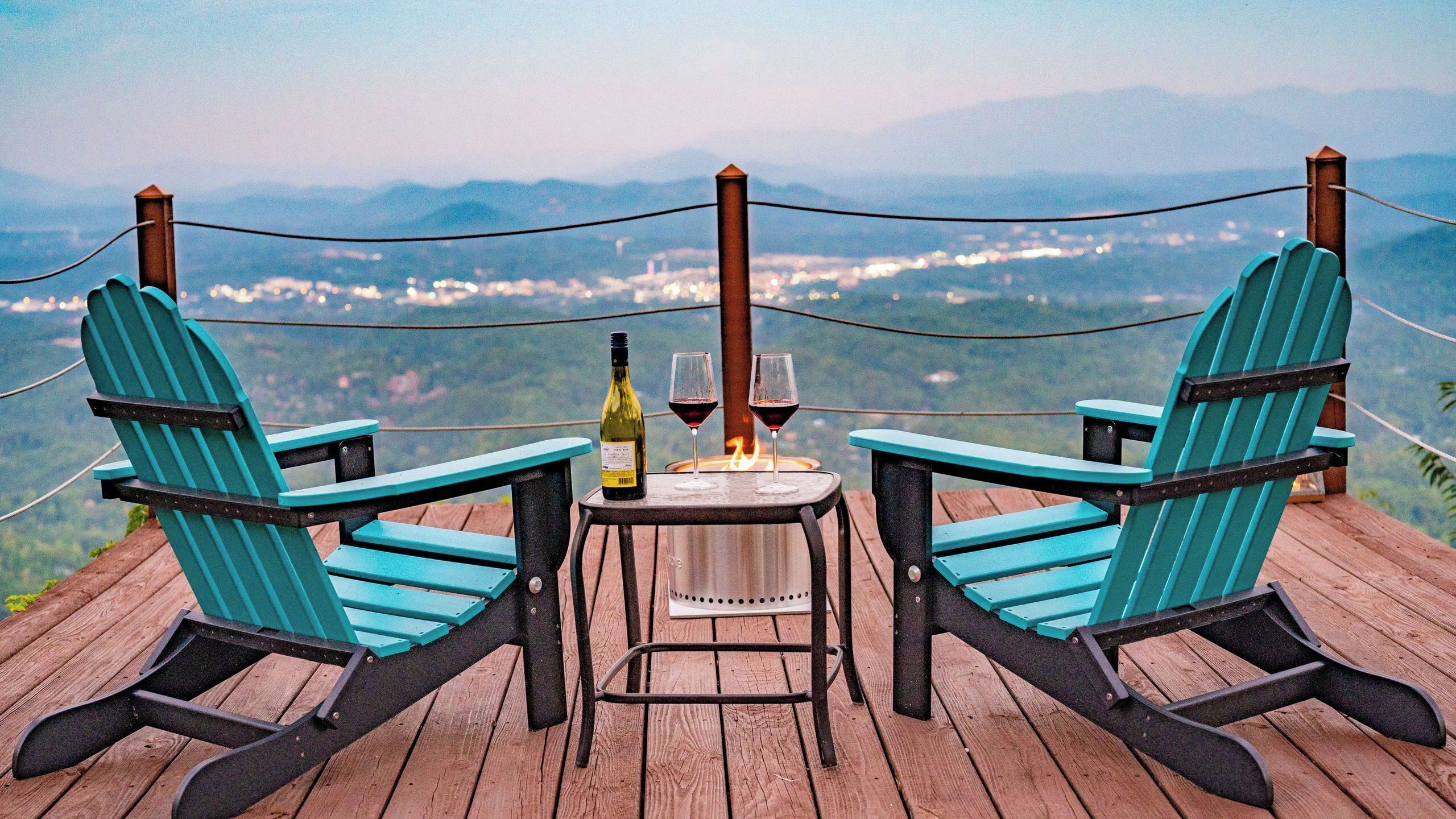 Zwei Adirondack-Stühle gegenüber, mit einer Weinflasche und zwei Gläsern auf einem kleinen Tisch, mit Blick auf eine malerische Aussicht auf eine Stadt und Berge.