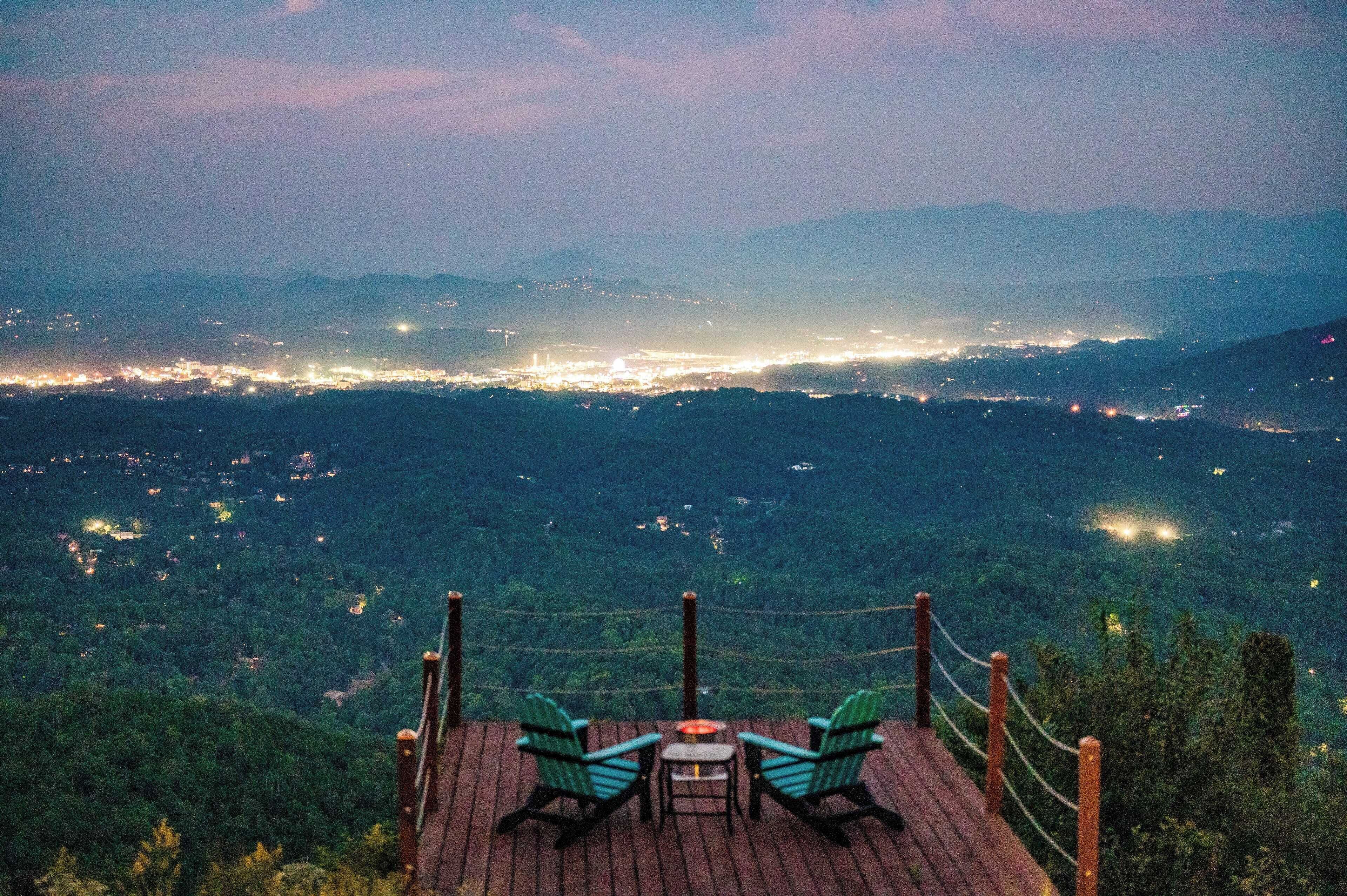 A high vantage point with a wooden deck, two chairs, and a table overlooking a cityscape at dusk.