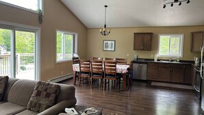 A spacious living room with a large window, a dining area with wooden chairs, a kitchen with wooden cabinets, and a ceiling fan. - 4 Bedroom Cottage- 129 Big Tub Rd (Tobermory)