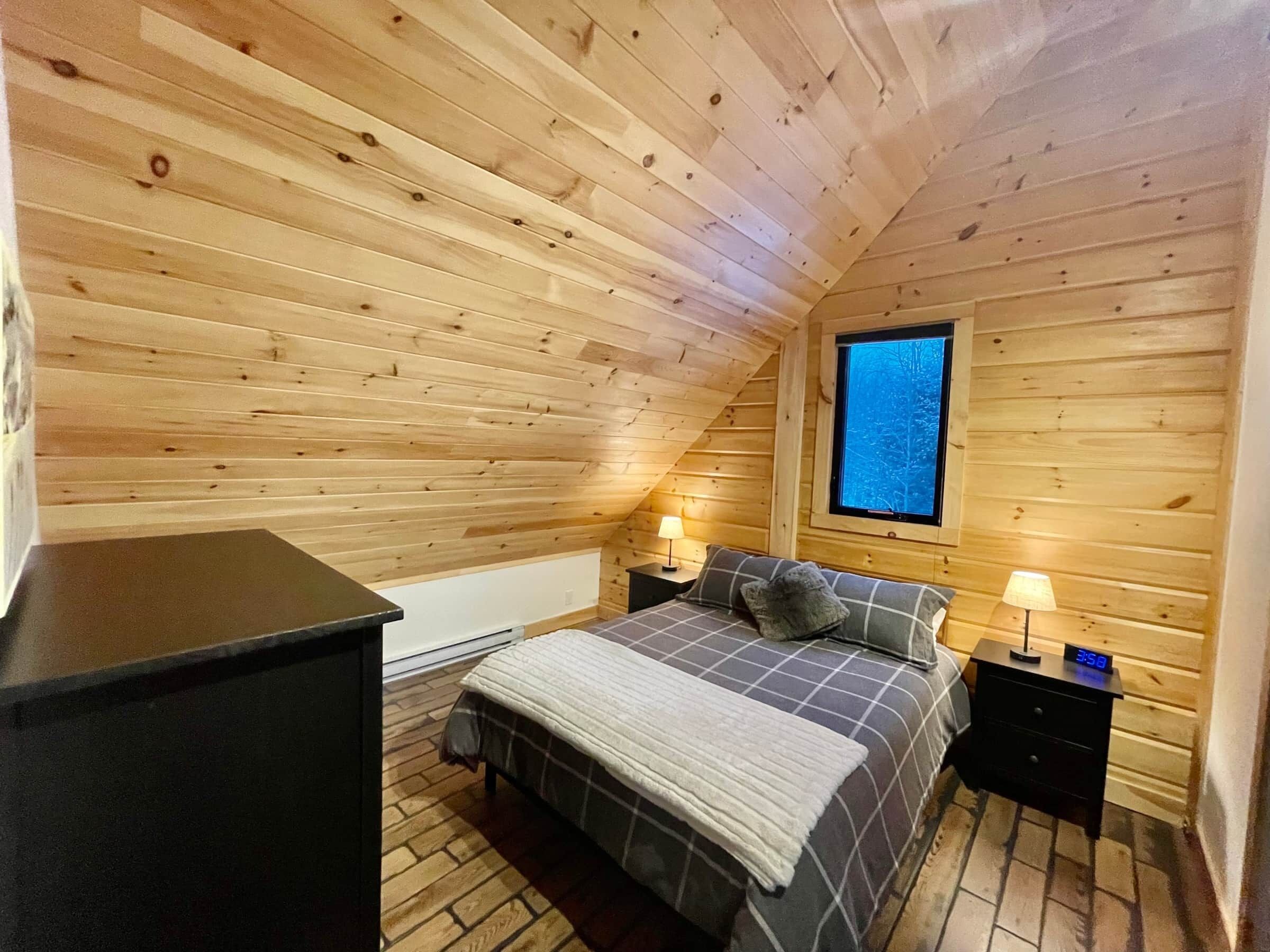 A cozy wooden cabin bedroom with a bed, two bedside tables, a nightstand, and a window with a view of trees.