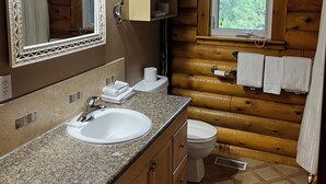Een houten badkamer van een chalet met een wastafel met granieten blad, een spiegel, een toilet en een raam met uitzicht op bomen.