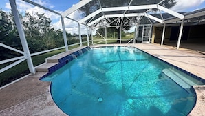 Ein Schwimmbecken mit klarem, blauem Wasser, umgeben von einer überdachten Fläche mit Metallgestell und Blick auf Bäume und Rasen.