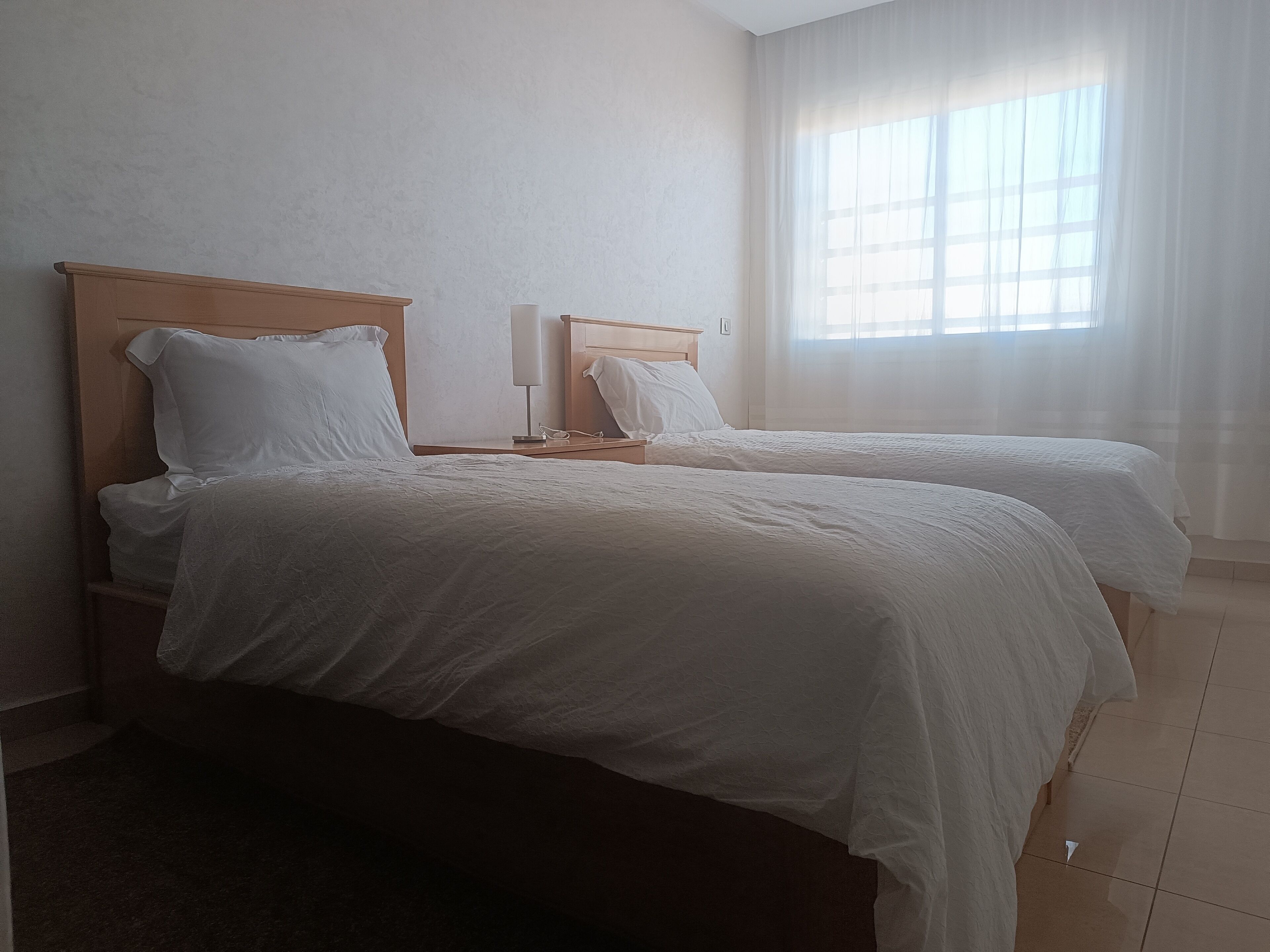 A hotel room with two single beds, a nightstand with a lamp, a window with sheer curtains, and a tiled floor.