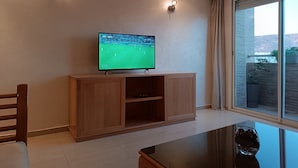 Ein modernes Wohnzimmer mit Flachbildfernseher an einem Holzkabinett, zwei Wandleuchten, einem Esstisch und einem großen Fenster mit Blick ins Freie.
