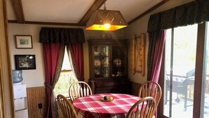 A dining area with a checkered tablecloth, wooden chairs, a water dispenser, a cabinet with glass doors, and a view of the outdoors through large windows.