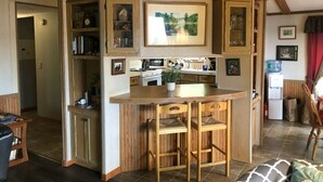 A compact kitchen with wooden cabinets, a central island with two stools, and a dining area with a chair.