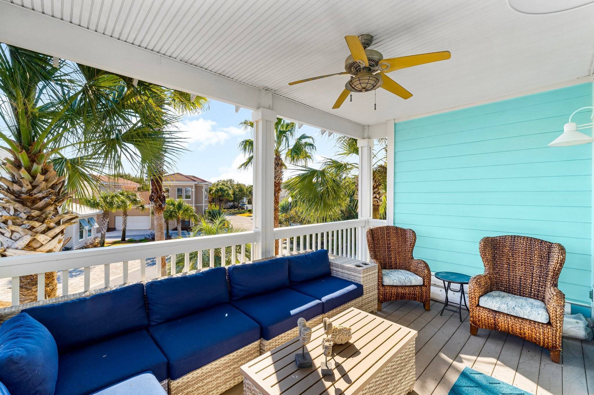 A covered porch with a blue wall, a ceiling fan, wicker furniture, and a view of palm trees and houses.