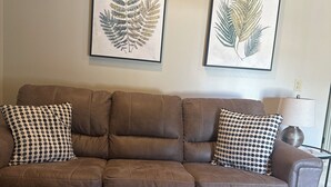 Ein braunes Ledersofa mit karierten Kissen, ein runder Couchtisch und zwei gerahmte botanische Drucke an der Wand.