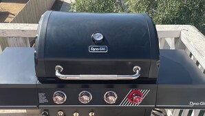 Ein schwarzer Grill für den Außenbereich mit dem Markennamen "Dyna-Glo", auf einem Balkon mit Holzgeländer und Grünpflanzen im Hintergrund.