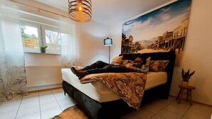 Un dormitorio con cama, ventana, radiador, televisor instalado en la pared y un mural de paisaje urbano.