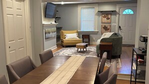 Un área de comedor con una mesa larga, sillas, ventilador de techo, luces empotradas, televisor, sofá y una ventana con persianas.
