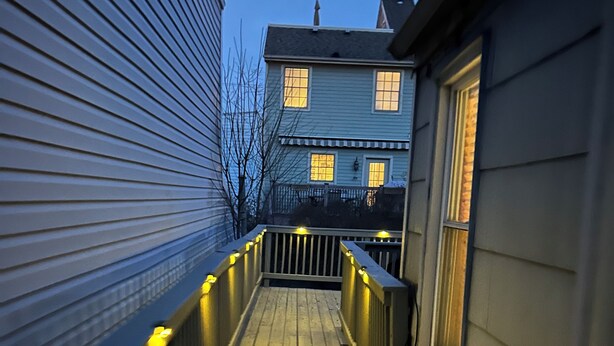Una terraza de madera con barandillas y luces colgantes, que conduce a una casa con múltiples ventanas.