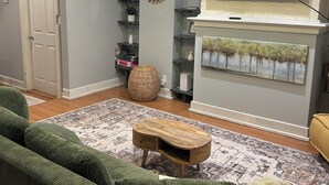 Una sala de estar con un sofá verde, una mesa de centro de madera, un televisor de pantalla plana sobre una chimenea y una alfombra estampada.