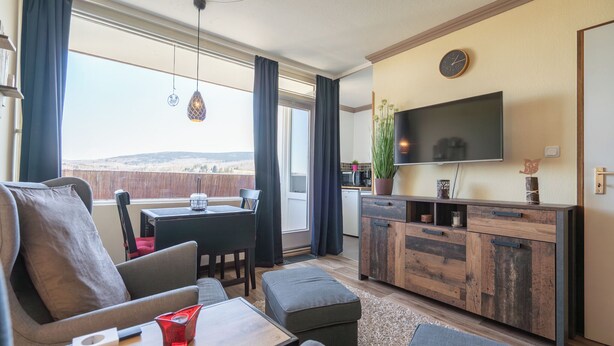 A modern living room with a sofa, ottoman, and coffee table, a dining area with a table and chairs, a kitchen area with a TV, and a view of the outdoors.