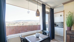 A balcony with a view of a hilly landscape, a dining table with chairs, a hanging pendant light, and a kitchen area visible through an open door.