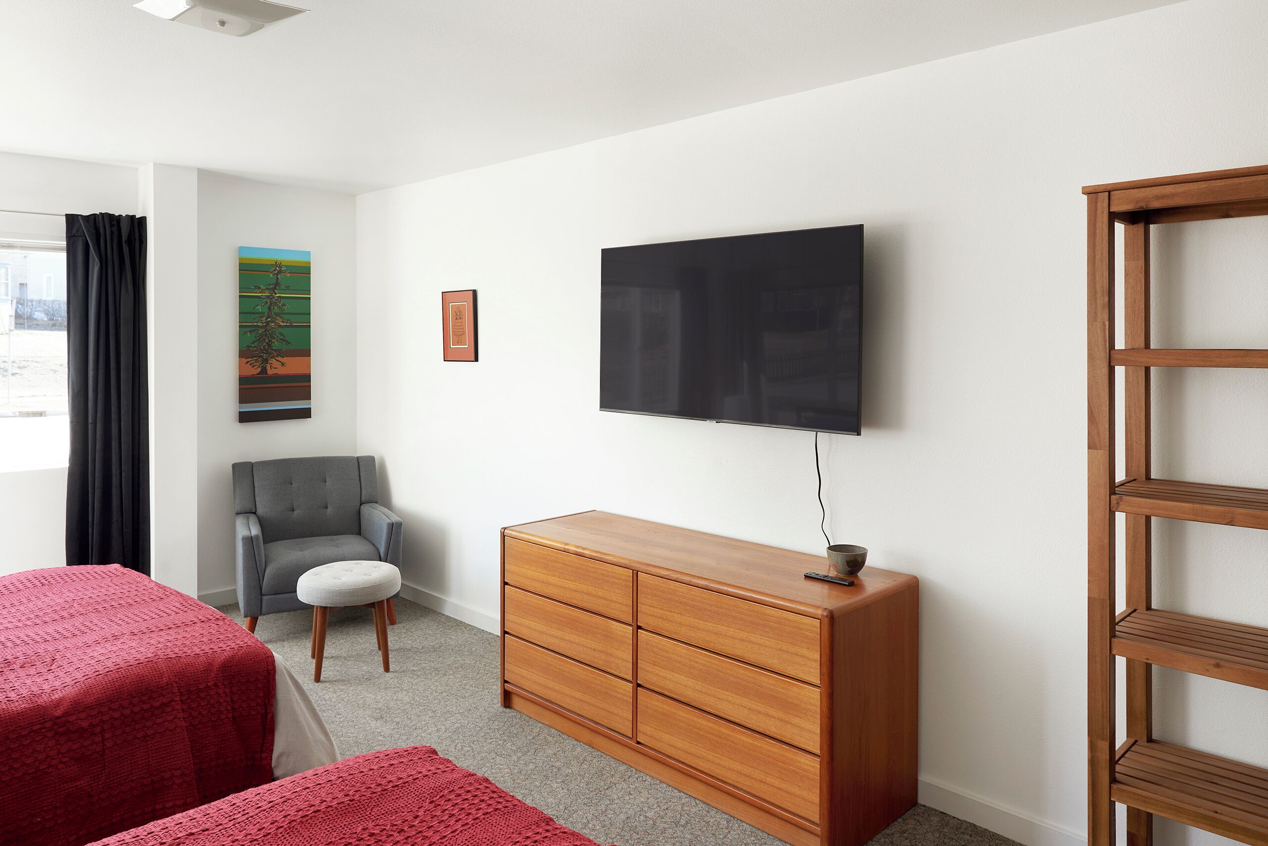 Een hotelkamer met een bed, een grijze fauteuil, een houten dressoir, een televisie en een raam met gordijnen.