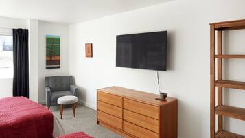Een hotelkamer met een bed, een grijze fauteuil, een houten dressoir, een televisie en een raam met gordijnen.