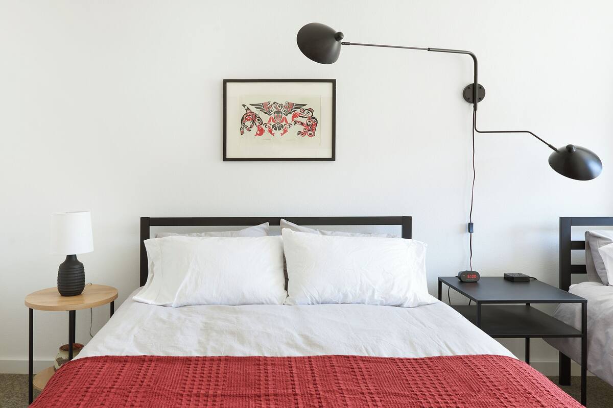 Un letto ben rifatto con biancheria bianca e rossa, una scrivania nera con una lampada e un'opera d'arte incorniciata appesa al muro.