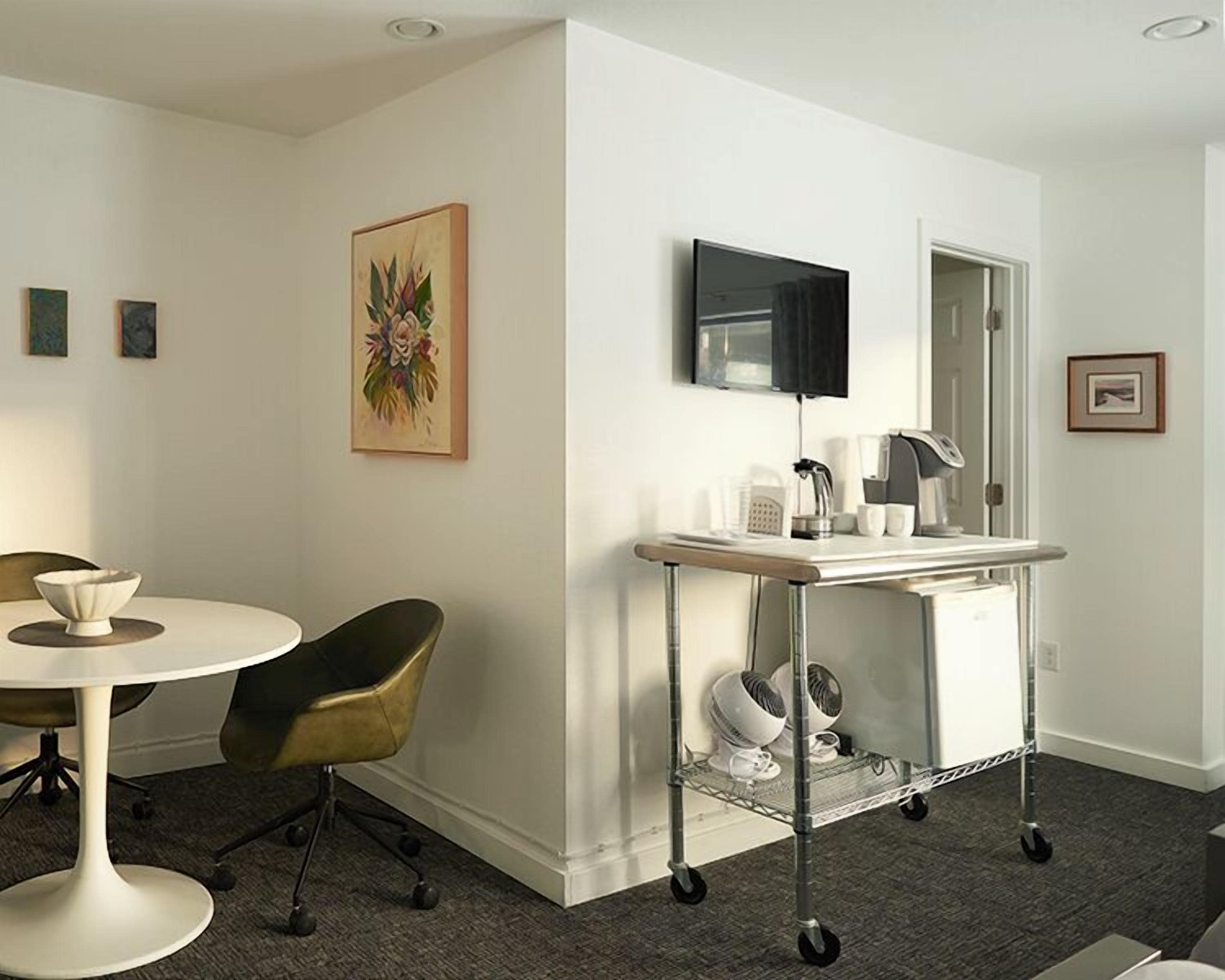Una habitación moderna con una mesa de comedor redonda, una silla de oficina negra, un escritorio blanco con un monitor de computadora, una máquina de café y un carrito con electrodomésticos.