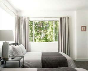 Un cuarto de hotel con un ventanal grande, una cama, una mesita de noche con una lámpara y un cuadro enmarcado en la pared.