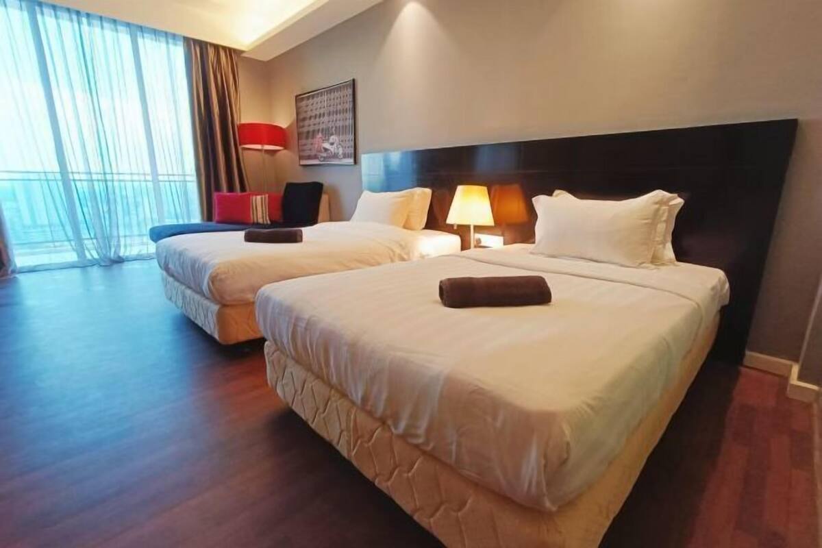 Habitación de hotel con dos camas, un ventanal con cortinas, una silla roja y una lámpara de pared.