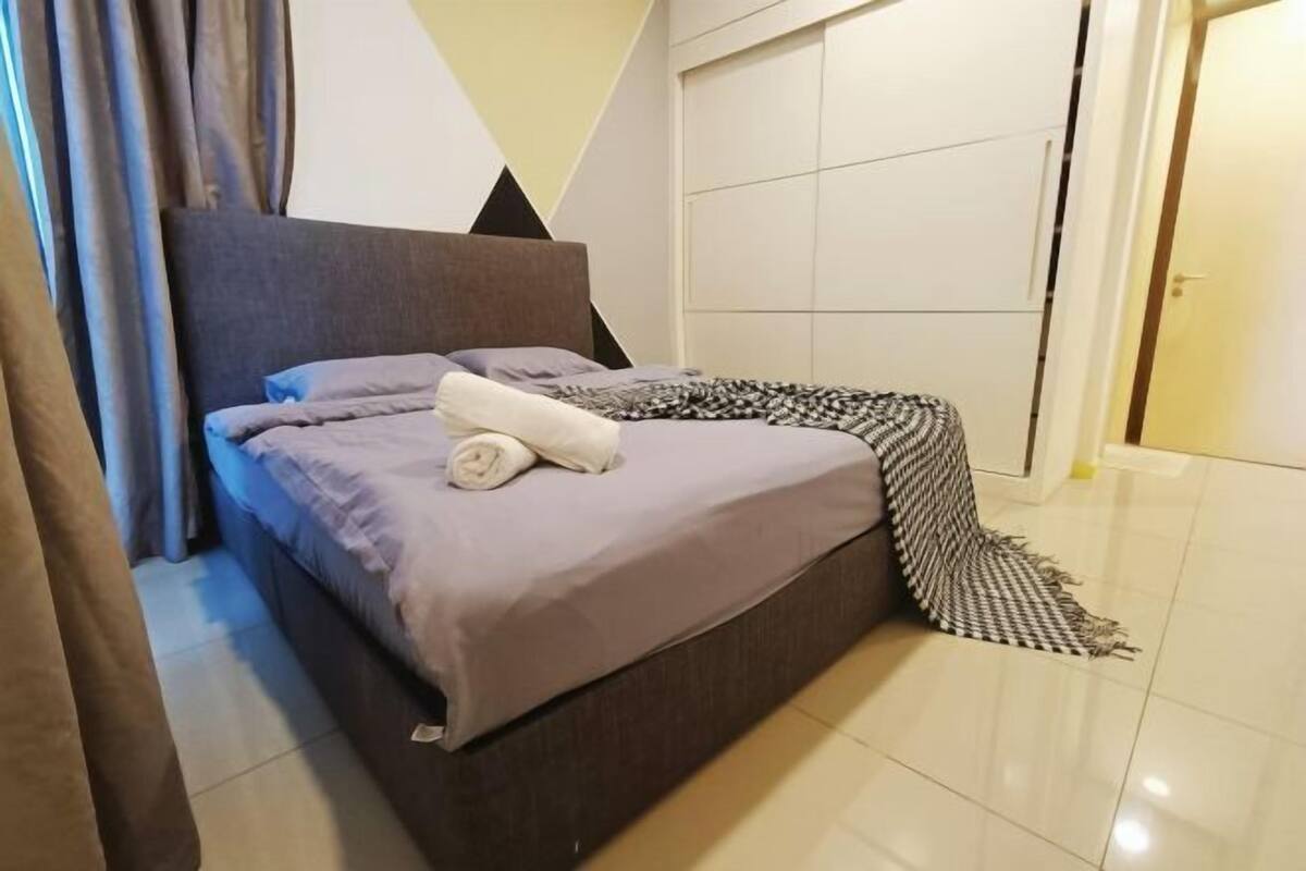 Una cama bien hecha con una manta gris y una toalla blanca enrollada, una manta decorativa estampada, y un diseño geométrico en la pared.