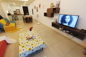 Una sala de estar con un sofá, una mesa de centro con mantel de flores, un televisor sobre un mueble bajo y una zona de comedor al fondo.