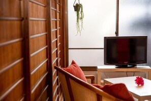 Een kamer met een televisie op een standaard, een houten stoel met een rode kussen en een klein tafeltje met een rode bloem.