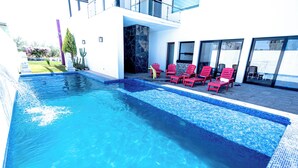 A swimming pool with clear blue water, surrounded by red lounge chairs and a modern building with large windows.