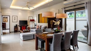 A modern living room with a dining area, a flat-screen TV, and a large window with curtains. - Emerald Pool Villa in Rawai (Phuket)