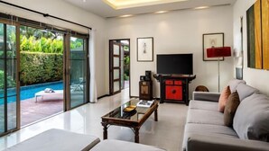 A modern living room with a flat-screen TV, a sofa, a coffee table, and a view of a swimming pool. - Emerald Pool Villa in Rawai (Phuket)