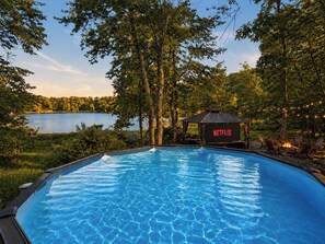 A swimming pool with a Netflix sign, surrounded by trees and a view of a lake. - Lakefront Chalet with Private Pool, Hot Tub, Sauna (East Stroudsburg)