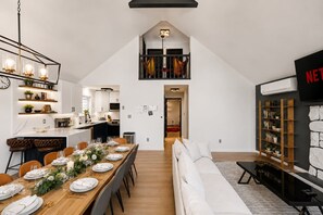 Une cuisine moderne avec des meubles blancs, un coin repas avec une table en bois dressée pour six personnes, un salon avec un canapé blanc et un escalier menant à une mezzanine.